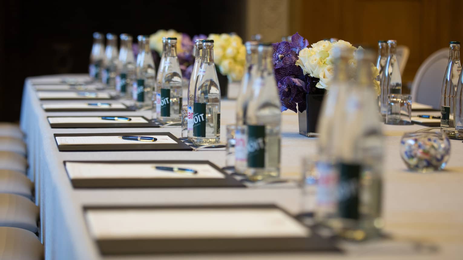Row of meeting agendas, glass bottles, small vases with flowers on boardroom table