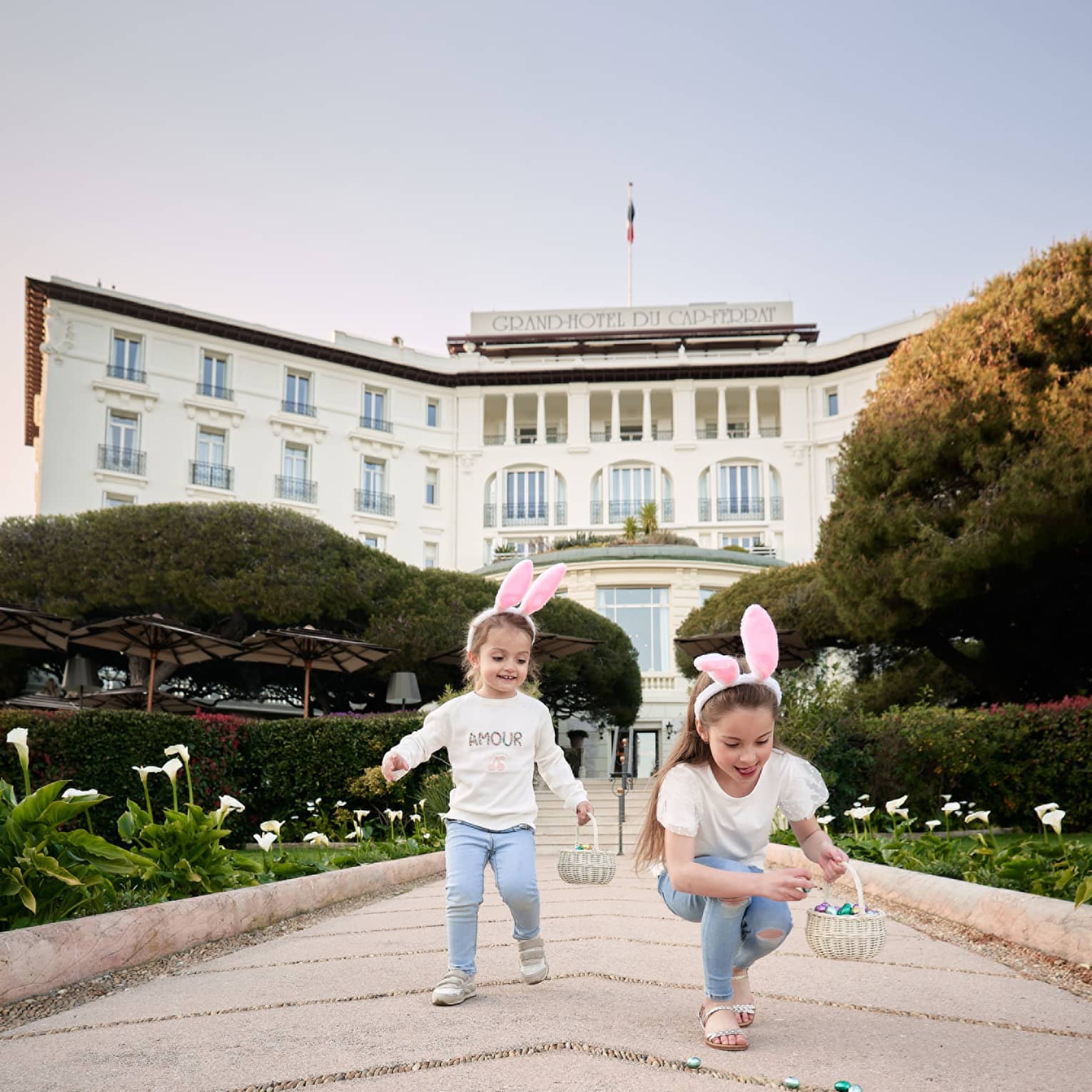 Two children in matching outfits wearing pink rabbit ears and holding Easter baskets with grand white building in backdrop