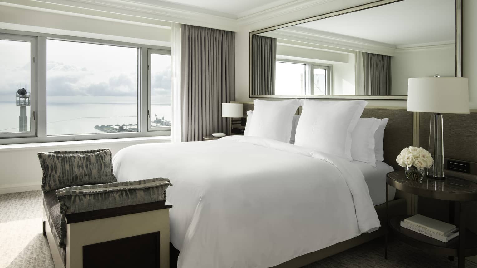 White bed, grey upholstered headboard, mirror above, nightstands with lamps. foot bench, window with lake view