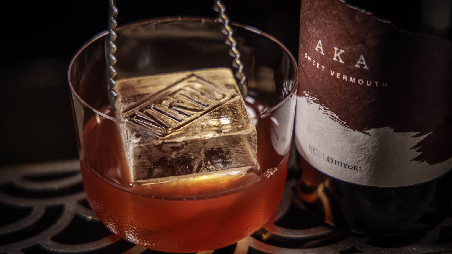 A pair of metal tongs lowers a large square ice cube into a chilled cocktail next to a bottle of Vermouth