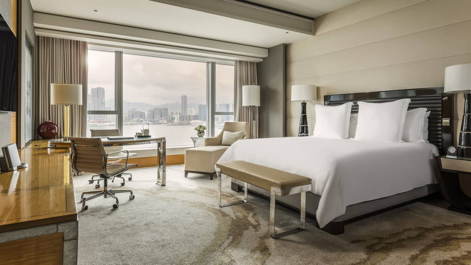 Deluxe Harbour-View Room bed, bench by desk, armchair at picture window overlooking city. harbour