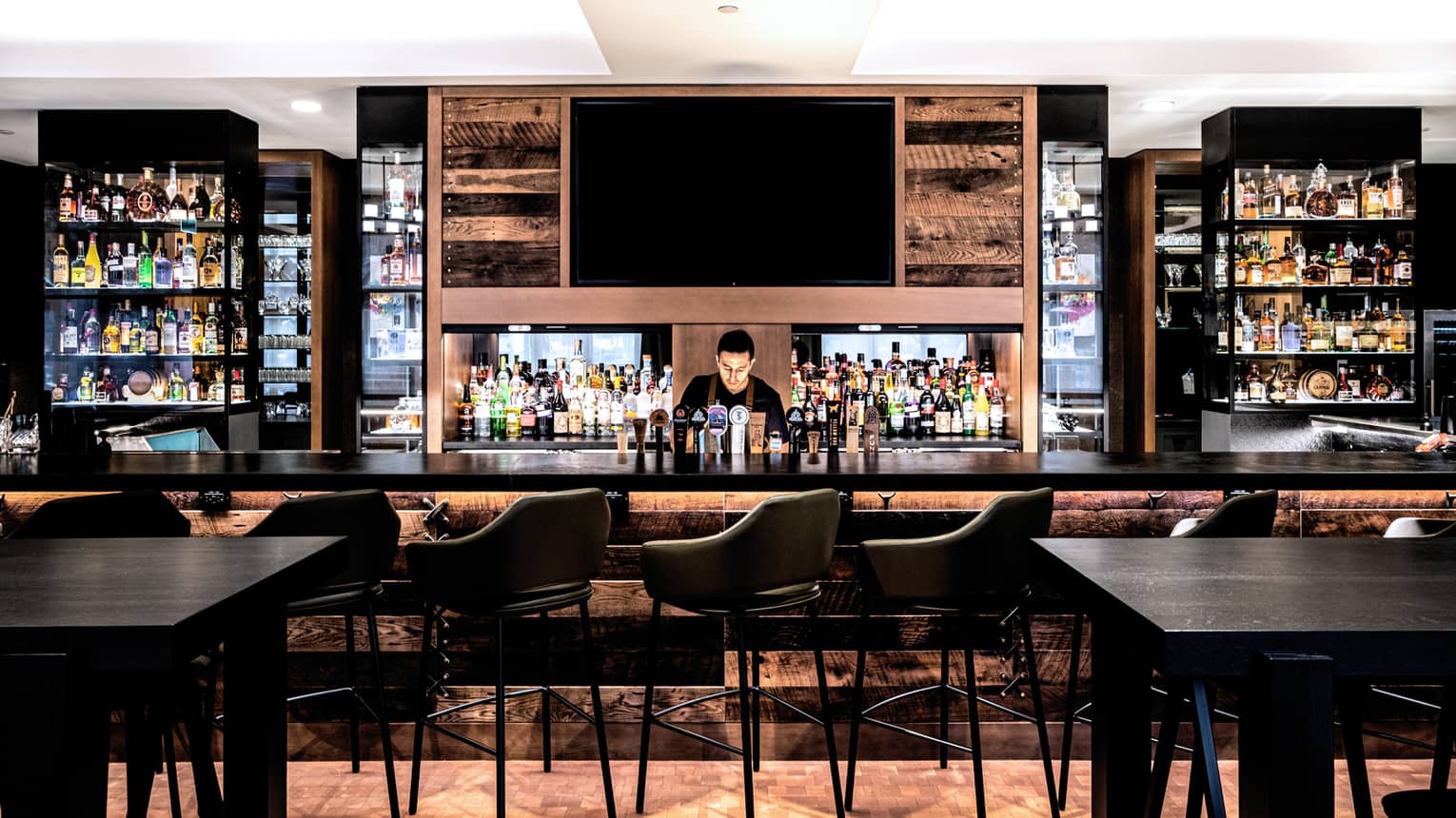A series of black barstools sit in front of a dimly lit bar with a single bartender.
