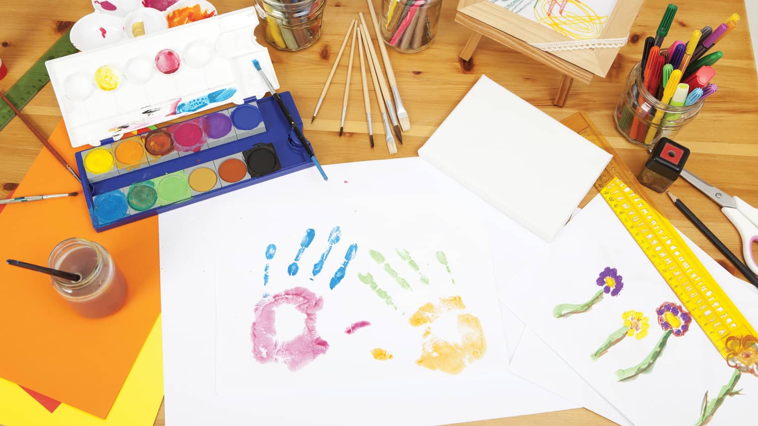 A creative workspace with watercolour paints, brushes, markers, handprint art, and drawings of flowers on a wooden table.