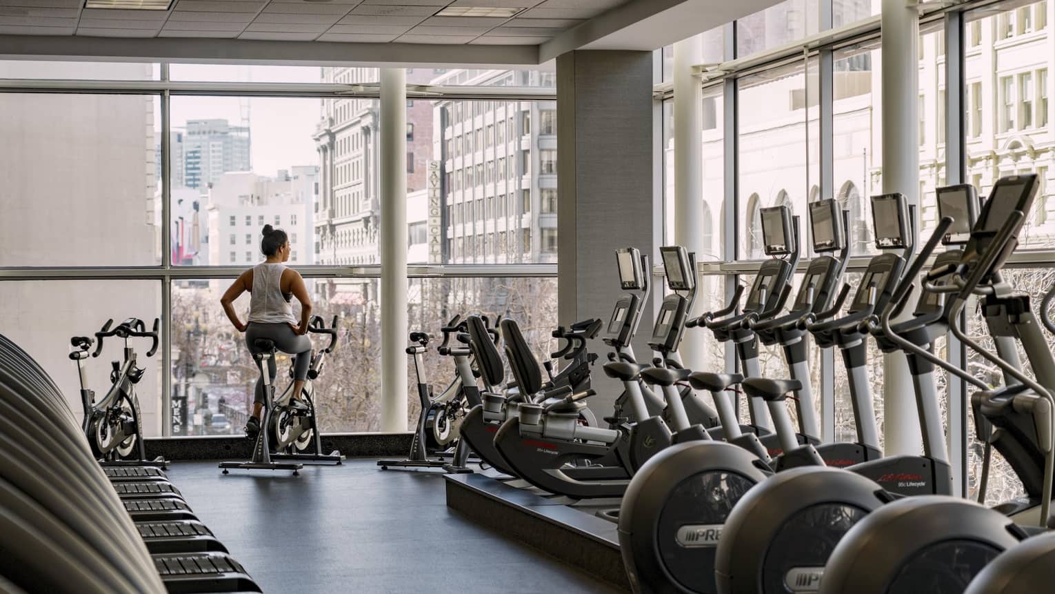 A woman rides a stationary bike in a hotel fitness center with floor-to-ceiling windows overlooking a city view