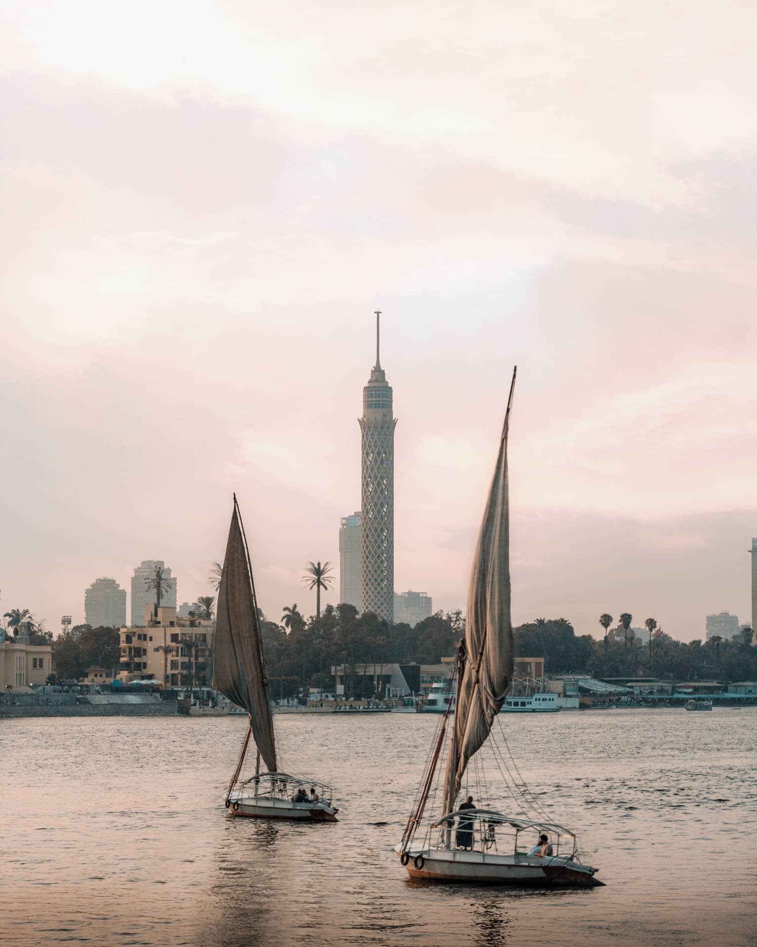 Two felucca boats drift on the water in the evening light, a busy shoreline and Cairo Tower in the distance. 