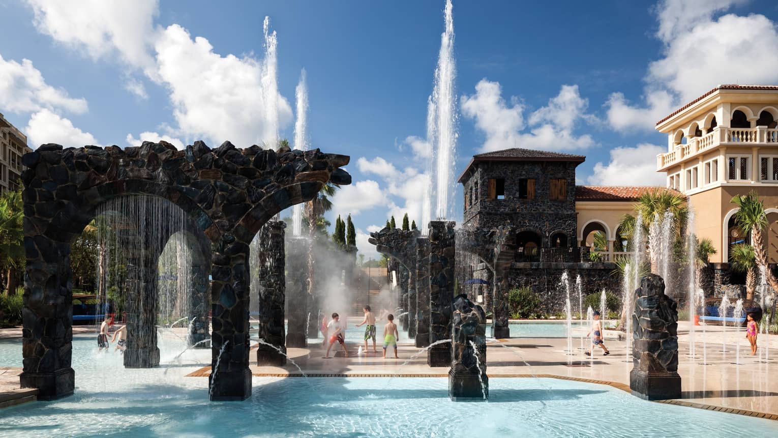 Kids play in water, under sprinklers and stone pillars in the Splash Zone water park