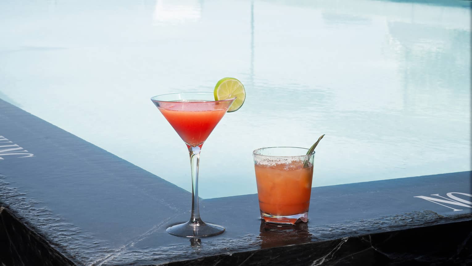A pink and orange cocktail on the edge of an outdoor pool.