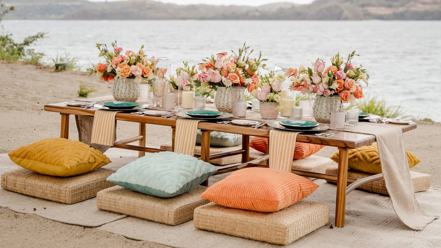 Beach dining set up with a low wodden table surrounded by colourful cushions set beneath a crochet umbrella