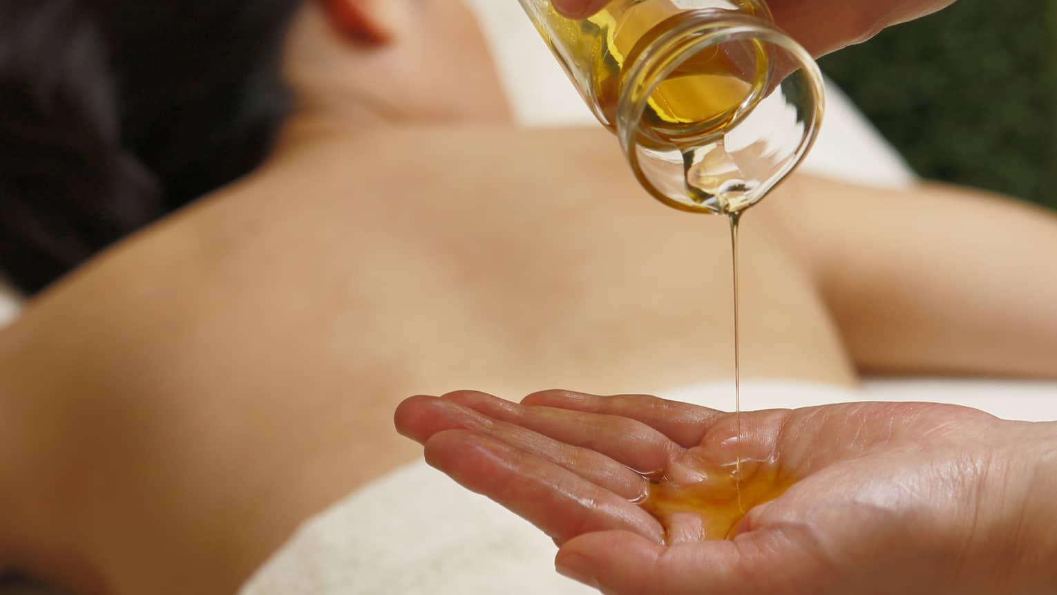 Spa staff pouring massage oil from glass jar in hand over woman lying on table