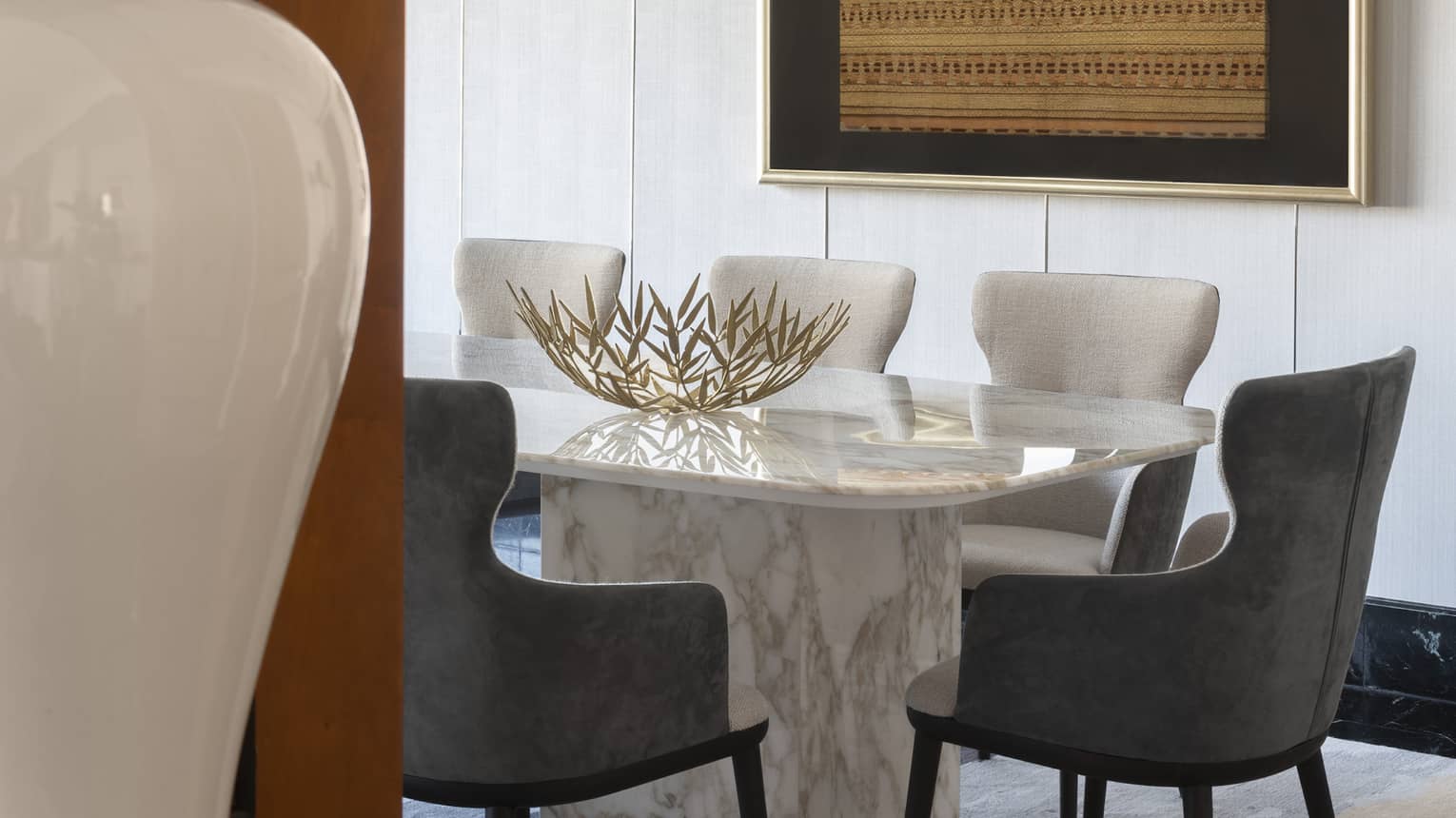 A modern dining area featuring a marble table, grey cushioned chairs and decorative centrepiece. The room is accented by framed artwork and a sleek chandelier.