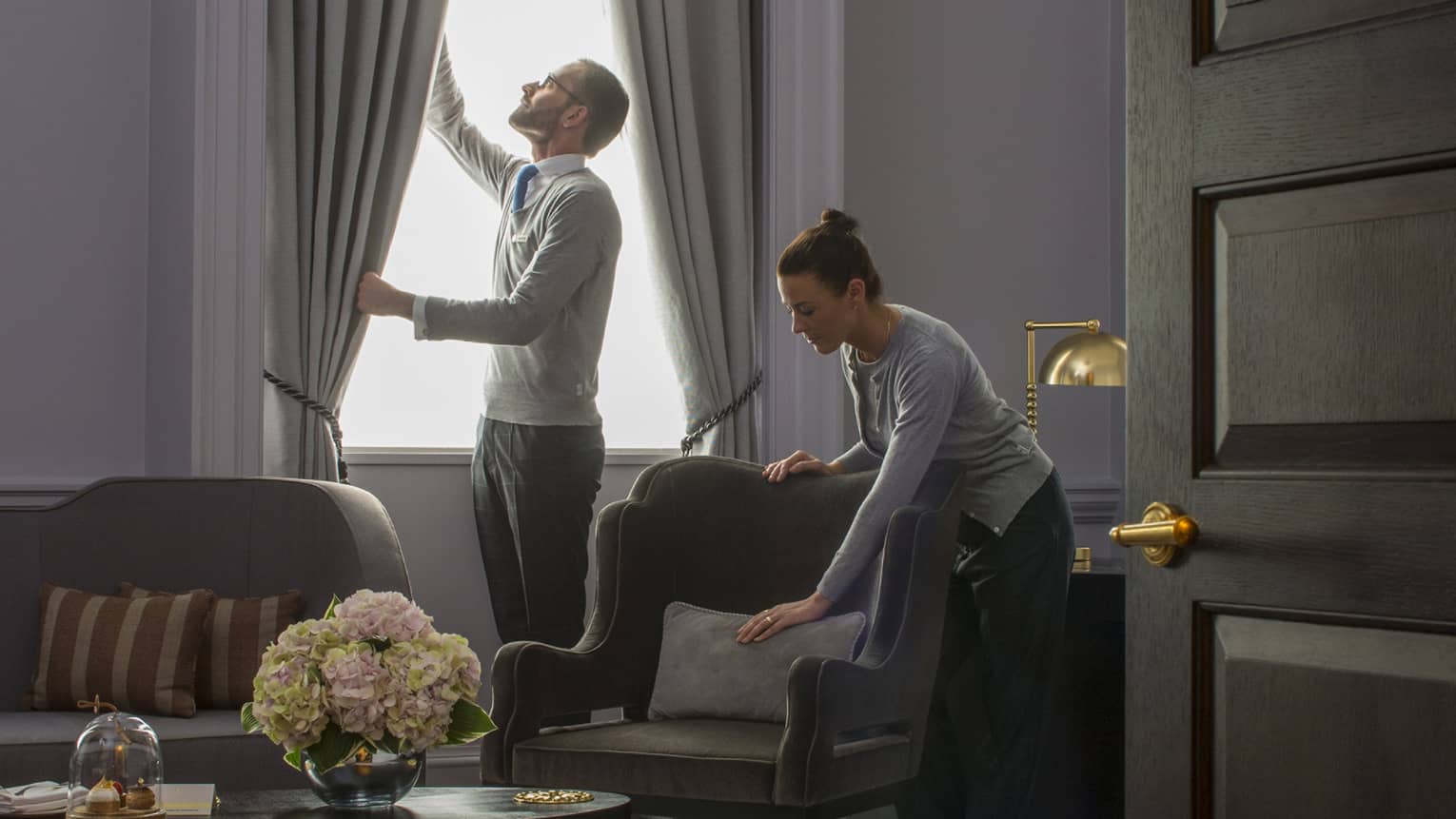 Grand Heritage Suite, man opens drapes as woman smooths pillow on armchair under chandelier 