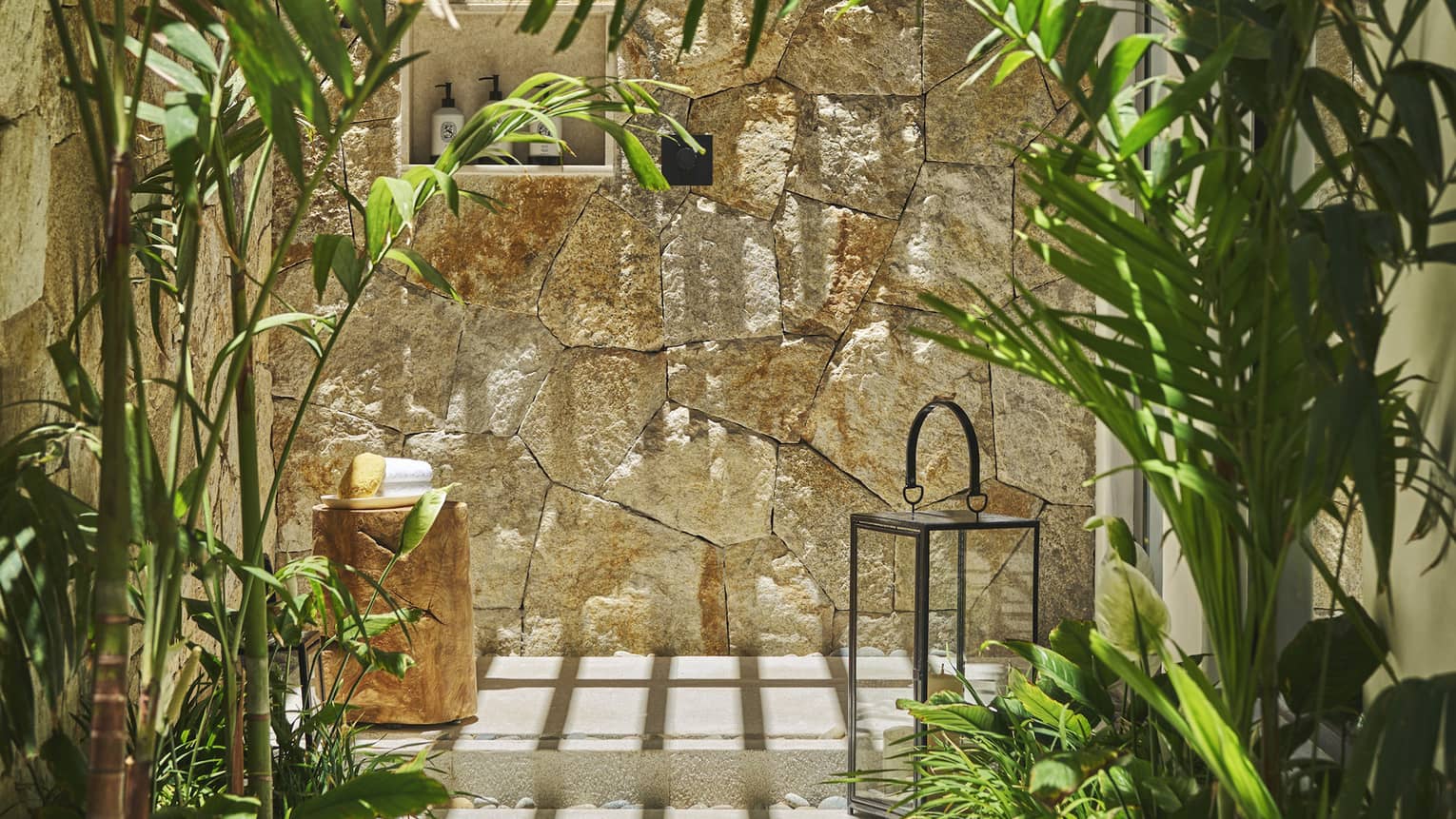 Casita outdoor shower area with a stone wall, surrounded by lush green plants, a wooden stool with towels and a decorative lantern