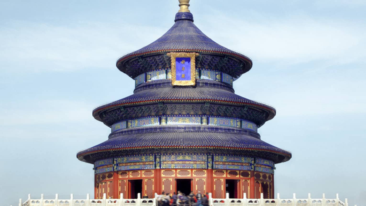 A close-up of a gilded, colourful triple-gabled circular temple bordered by a low, white balustrade against a clear blue sky.