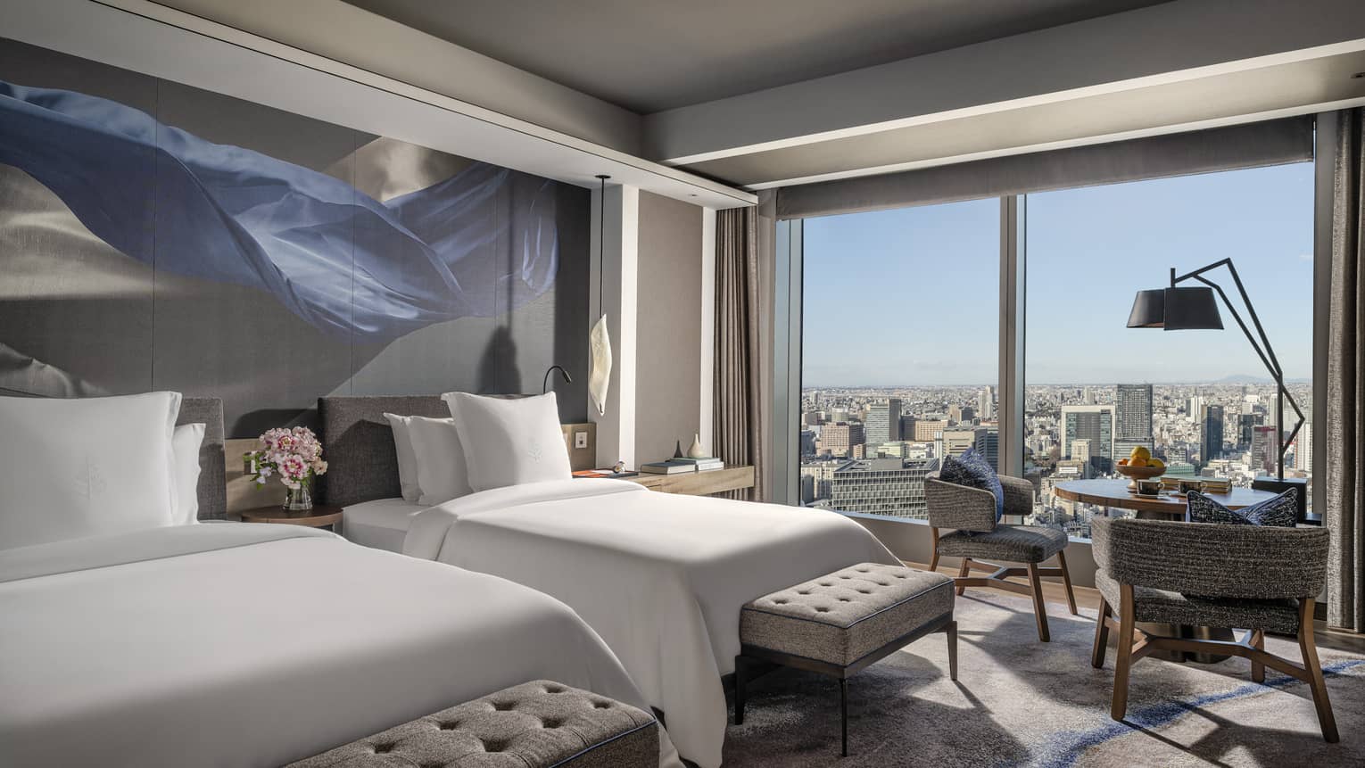 Hotel room with two single beds, small table and two chairs, and wall of windows looking out to city