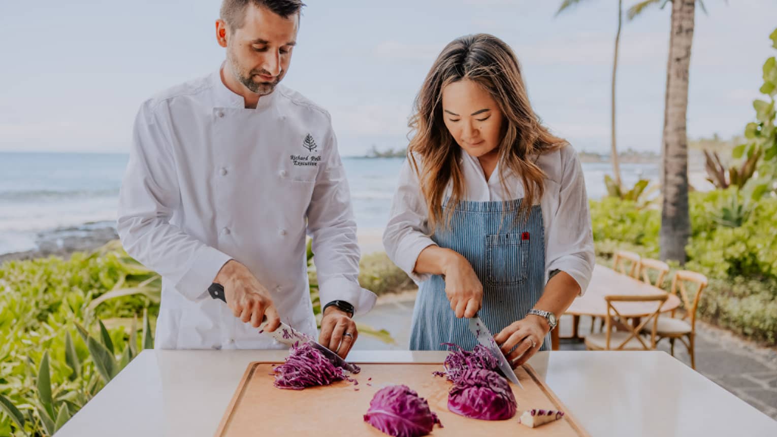 A woman and a man in a chef's jacket practice thinly slicing vegetables