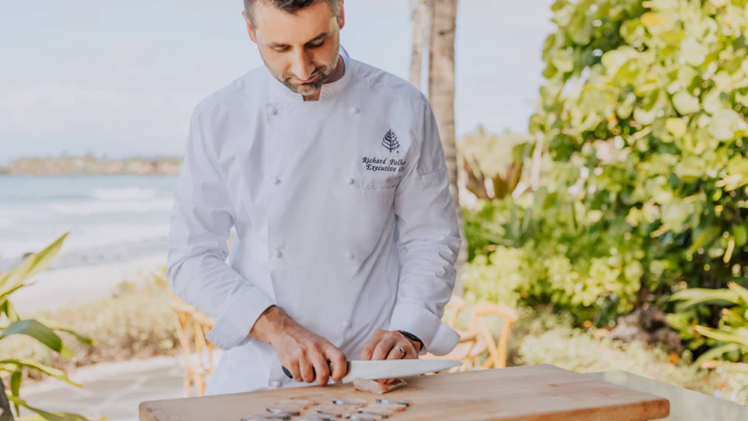 A male chef thinly slices seafood on wooden cutting board, beach in the background