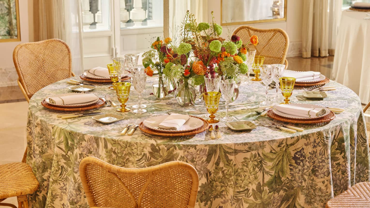 A well-lit interior event space with neutral decor features a round table with chairs and a floral tablecloth. The table is set with plates, napkins, utensils, clear and translucent gold stemware, small square side plates, and a colourful floral centerpiece.