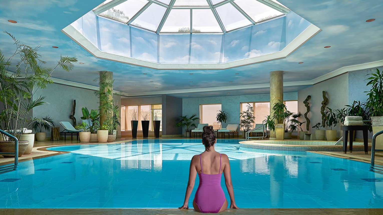 Woman in swimsuit seated on edge of indoor pool with blue ceiling and skylight above