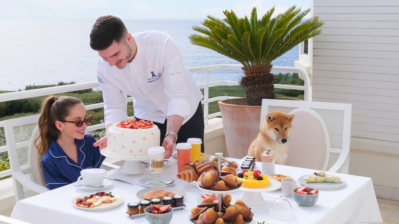 Chef presenting a white cake to child having breakfast with her dog at terrace table overlooking the sea