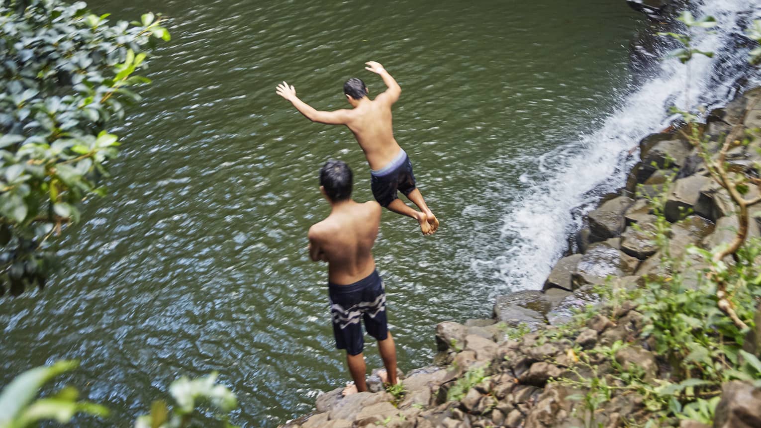 Two people leap from rocks to water below waterfall on the road to Hana