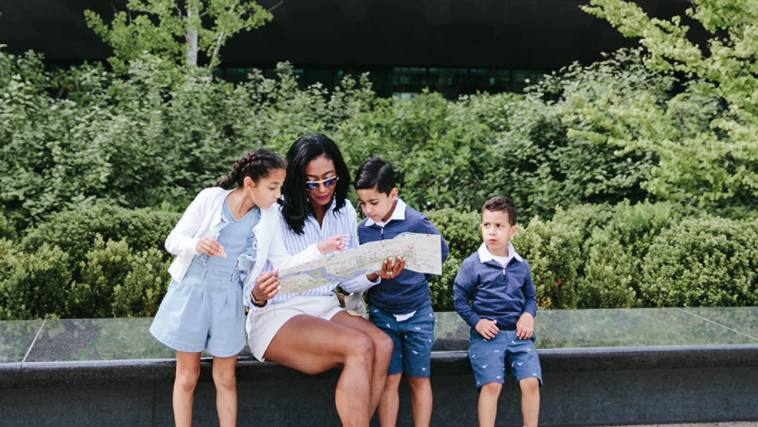 Woman and three young children sit on bench in city with landscaping behind them