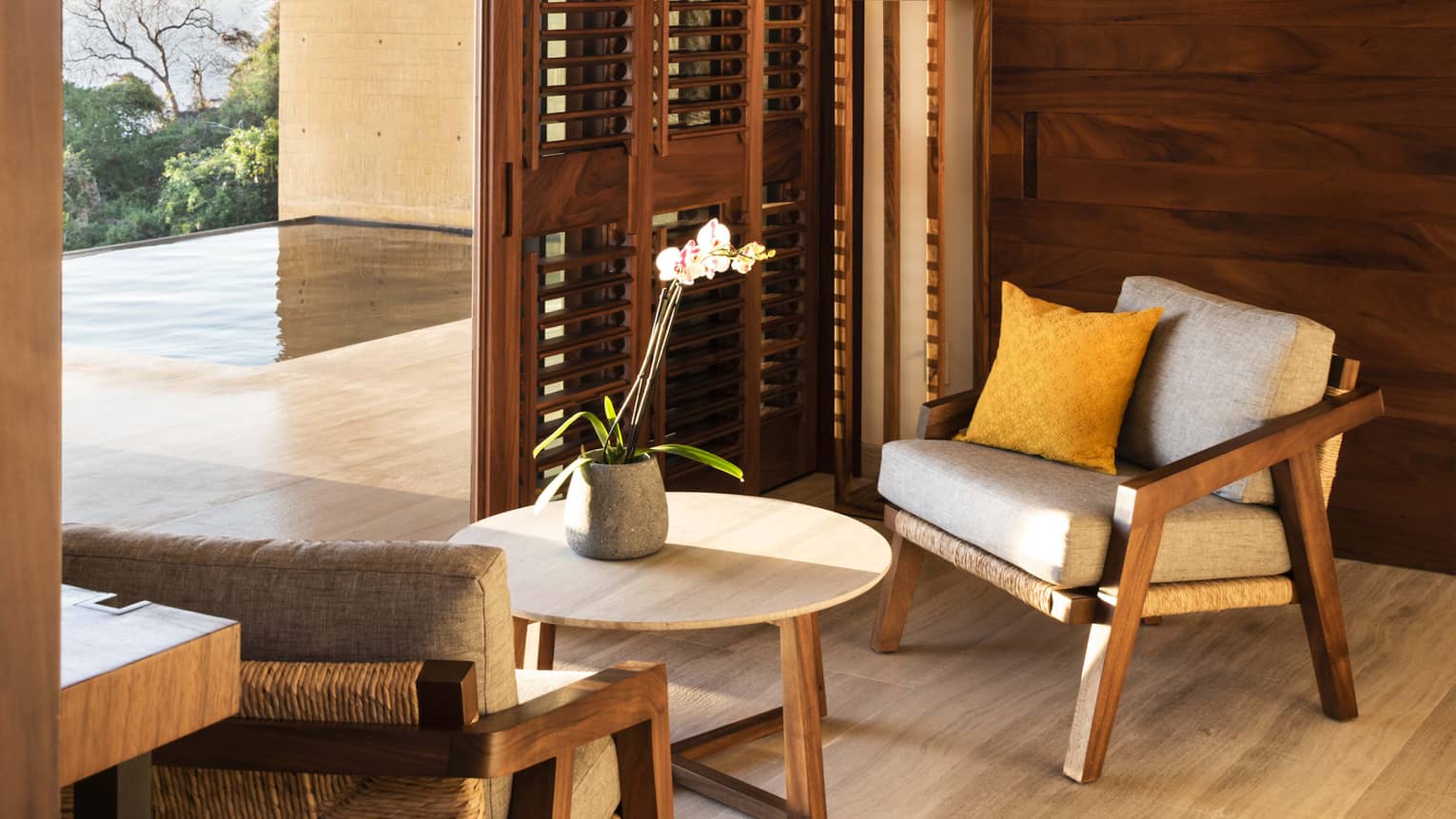 Cozy indoor seating area with wooden accents, featuring a pair of cushioned chairs and a small table. Large shutters open to a terrace with an infinity pool and garden views.