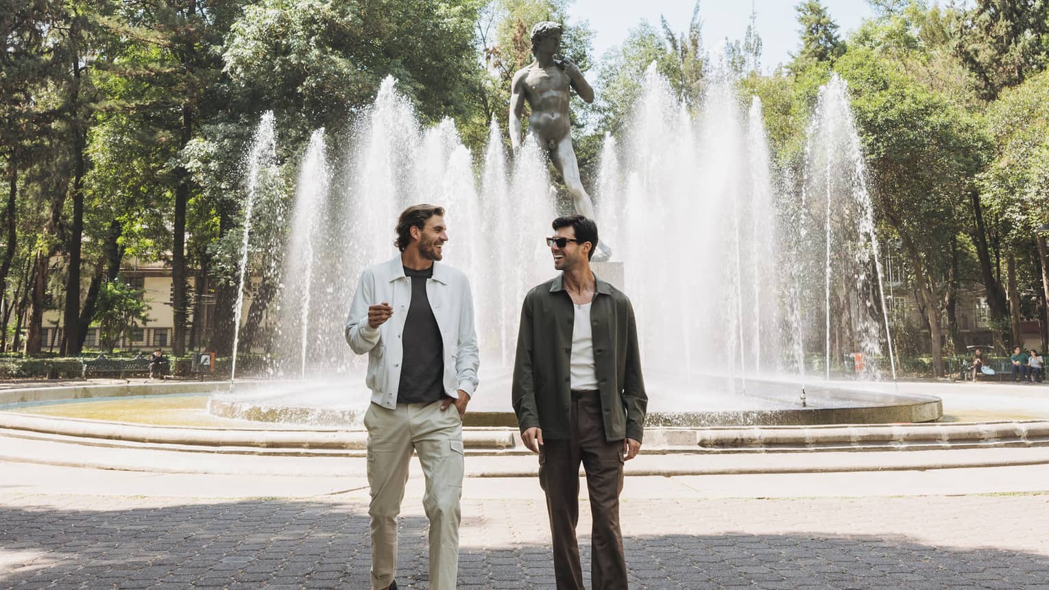 A pair in a sunny park chats in front of a Michelangelo's David statue surrounded by water jetting up from ground spouts. 
