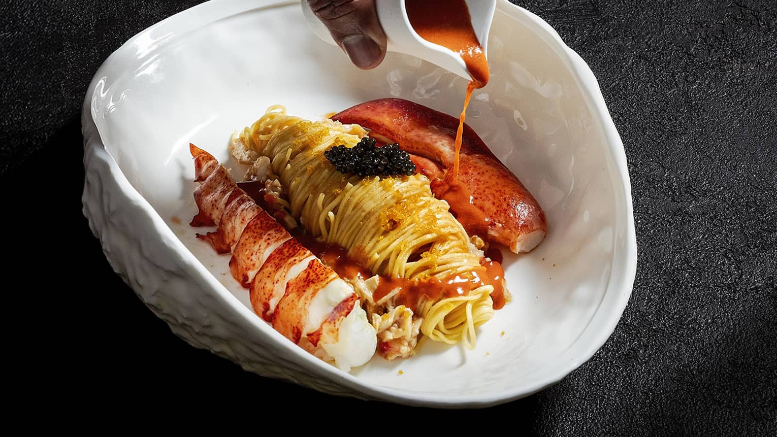 Hand pours red sauce from a white ceramic container onto a while bowl of pasta and seafood