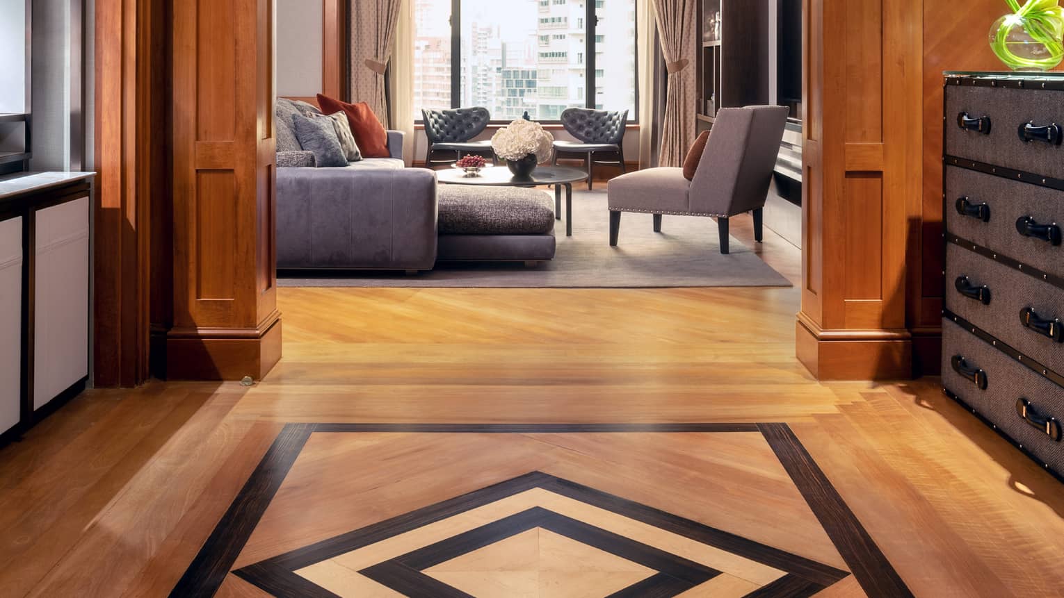 A warm, elegant hallway with wooden panelling and patterned wood floors leading to a living area with a sectional sofa, armchairs and large windows offering city views.