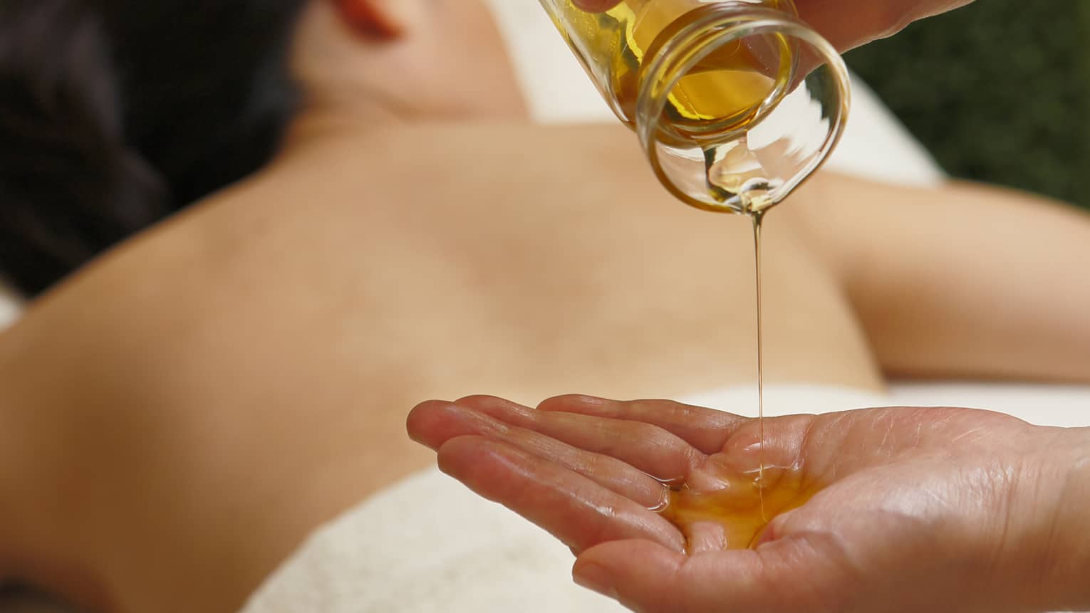 Spa staff pouring massage oil from glass jar in hand over woman lying on table