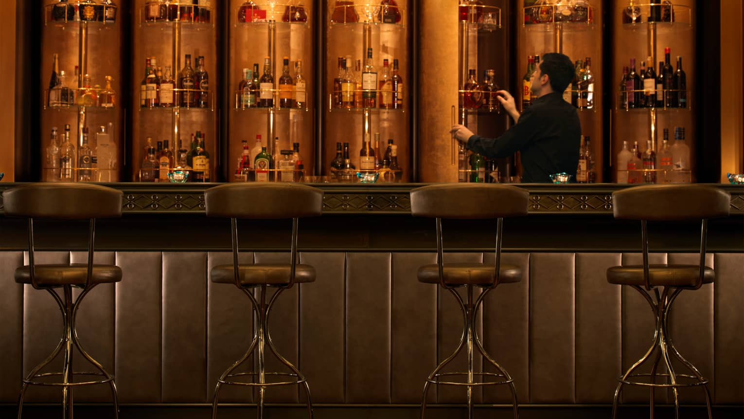 Bentleys dimly-lit lounge, four stools at padded leather bar where bartender looks at racks of bottles on wall