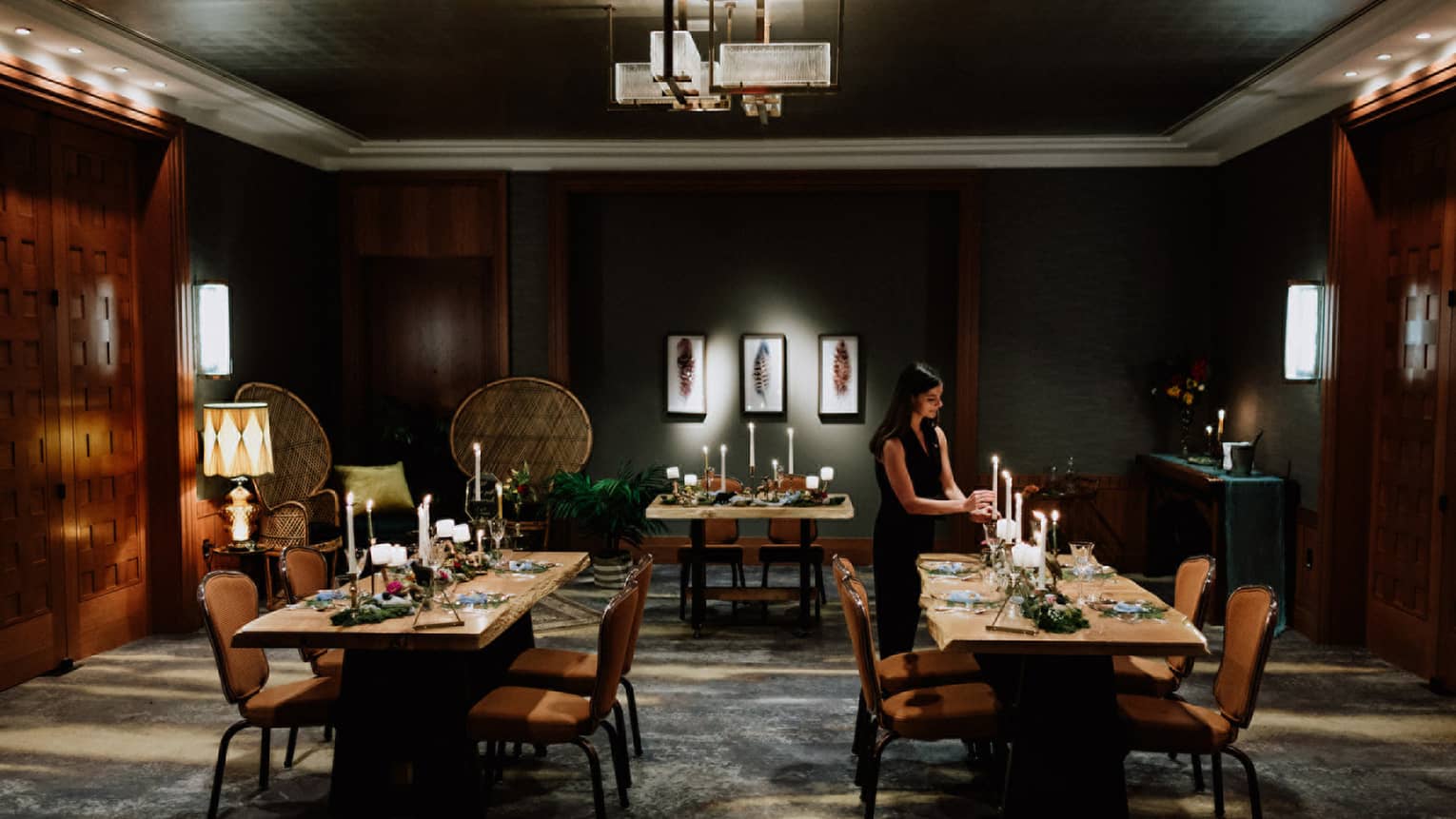 Intimate wedding dinner setup in a luxurious dining room at Four Seasons Whistler. The room features dark wood paneling and soft lighting, creating a warm and inviting atmosphere. Two wooden tables are elegantly set with white candles, floral arrangements, and fine dinnerware. A woman in a dark dress adjusts the table settings, preparing for the event. The sophisticated decor and thoughtful arrangement reflect a serene and exclusive wedding venue.,Intimate wedding dinner setup in a luxurious dining room at Four Seasons Whistler. The room features dark wood paneling and soft lighting, creating a warm and inviting atmosphere. Two wooden tables are elegantly set with white candles, floral arrangements, and fine dinnerware. A woman in a dark dress adjusts the table settings, preparing for the event. The sophisticated decor and thoughtful arrangement reflect a serene and exclusive wedding venue.