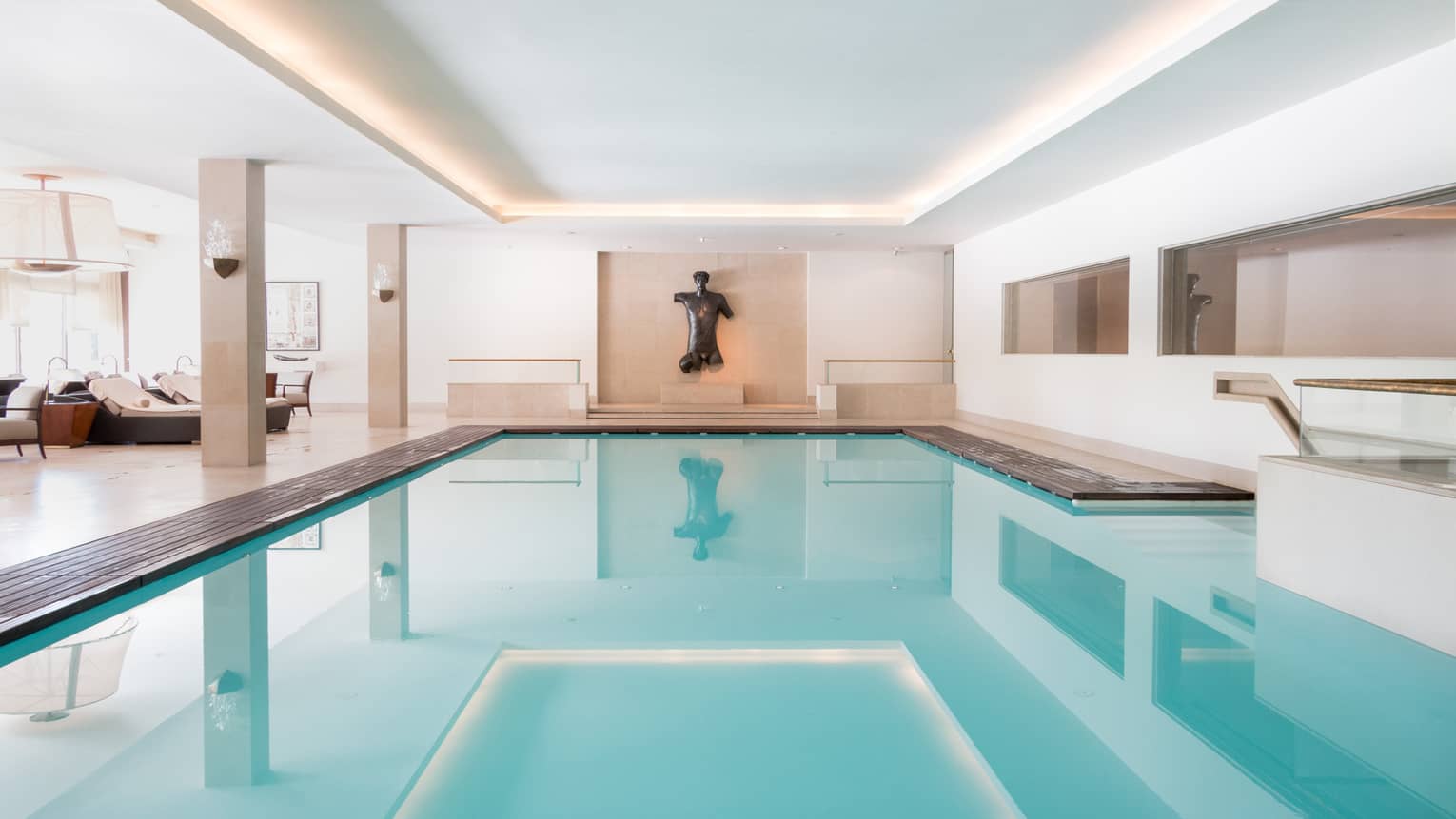 Rectangular pool with still blue water and flanked by lounge chairs ends at a wall adorned with hanging sculpture