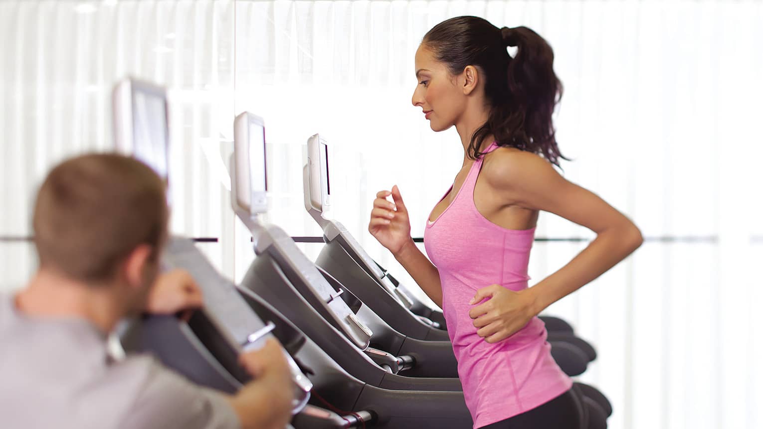 Woman in pink sleeveless shirt and ponytail runs on treadmill as personal trainer watches