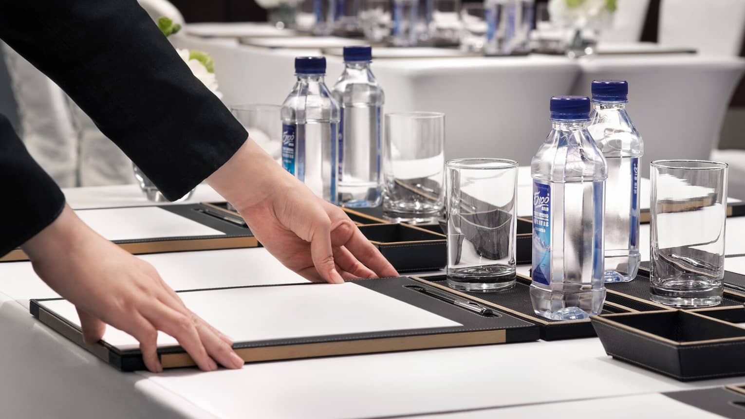 A person adjusts a writing pad on a conference room table with bottled water, glasses and floral arrangements