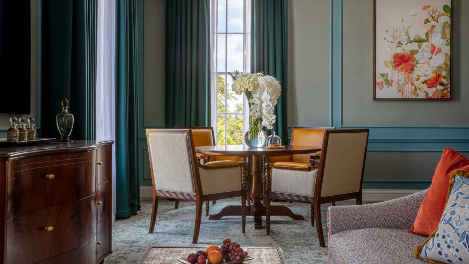 Guest room with round wooden table and four dining chairs, patterned couch, cherry sideboard, coffee table, windows with blue drapes