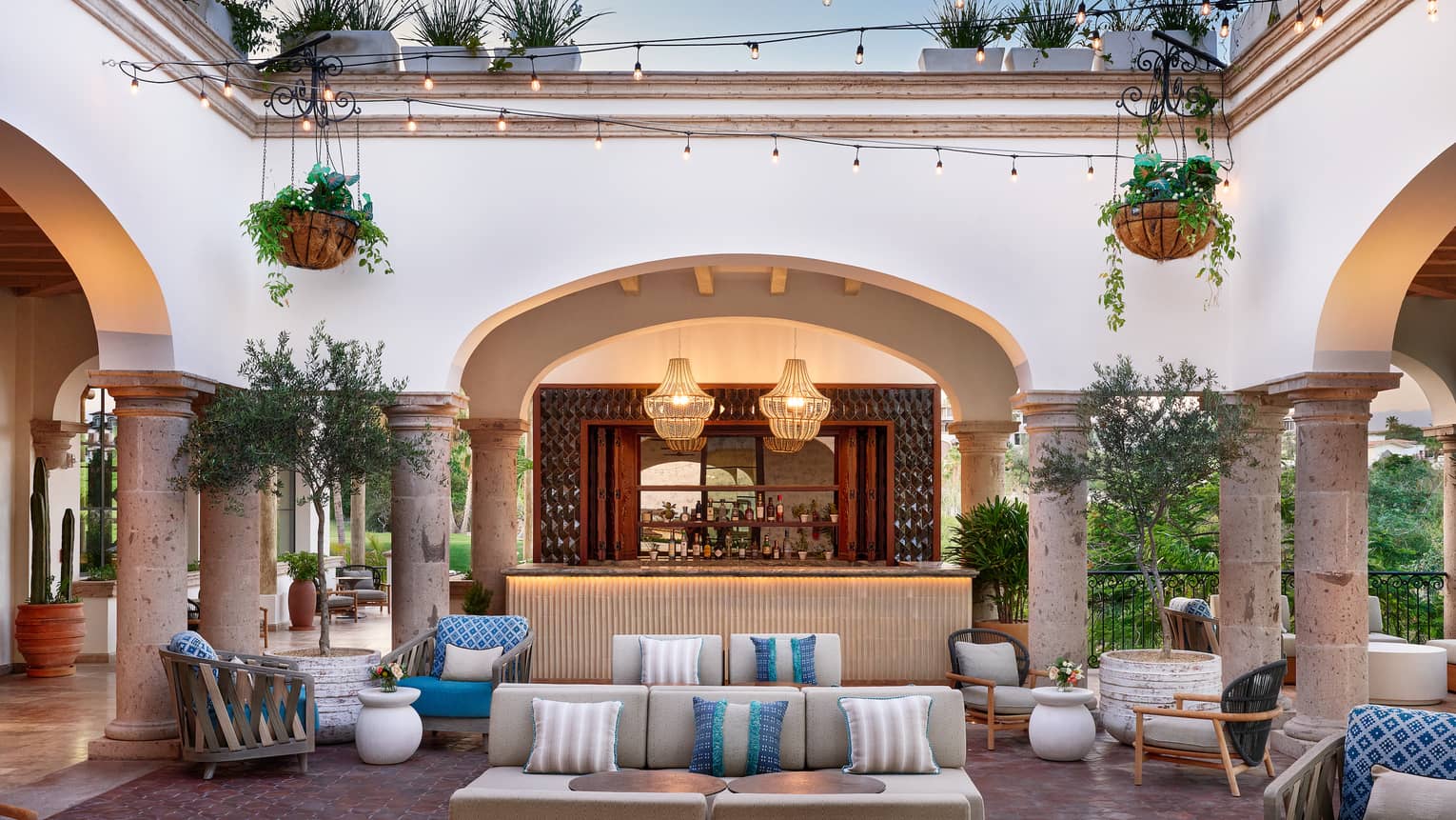 A spacious courtyard surrounded by arched openings. The space contains upholstered couches and chairs, along with potted and hanging plants, and string and pendant lighting.