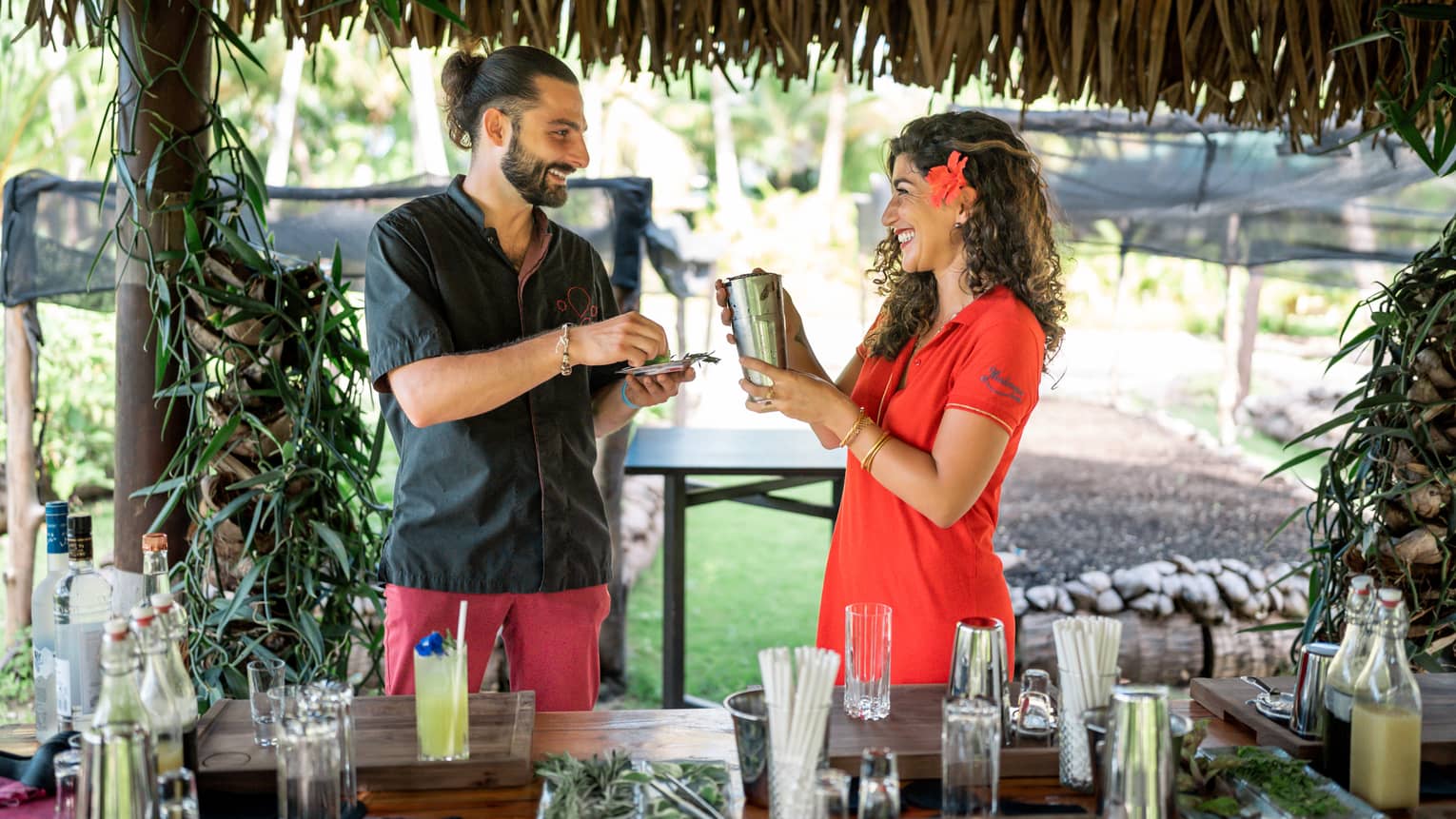 Two people hold a shaker and fresh herbs in a straw-roofed hut over a bar strewn with glasses, liquor bottles and garnishes. 