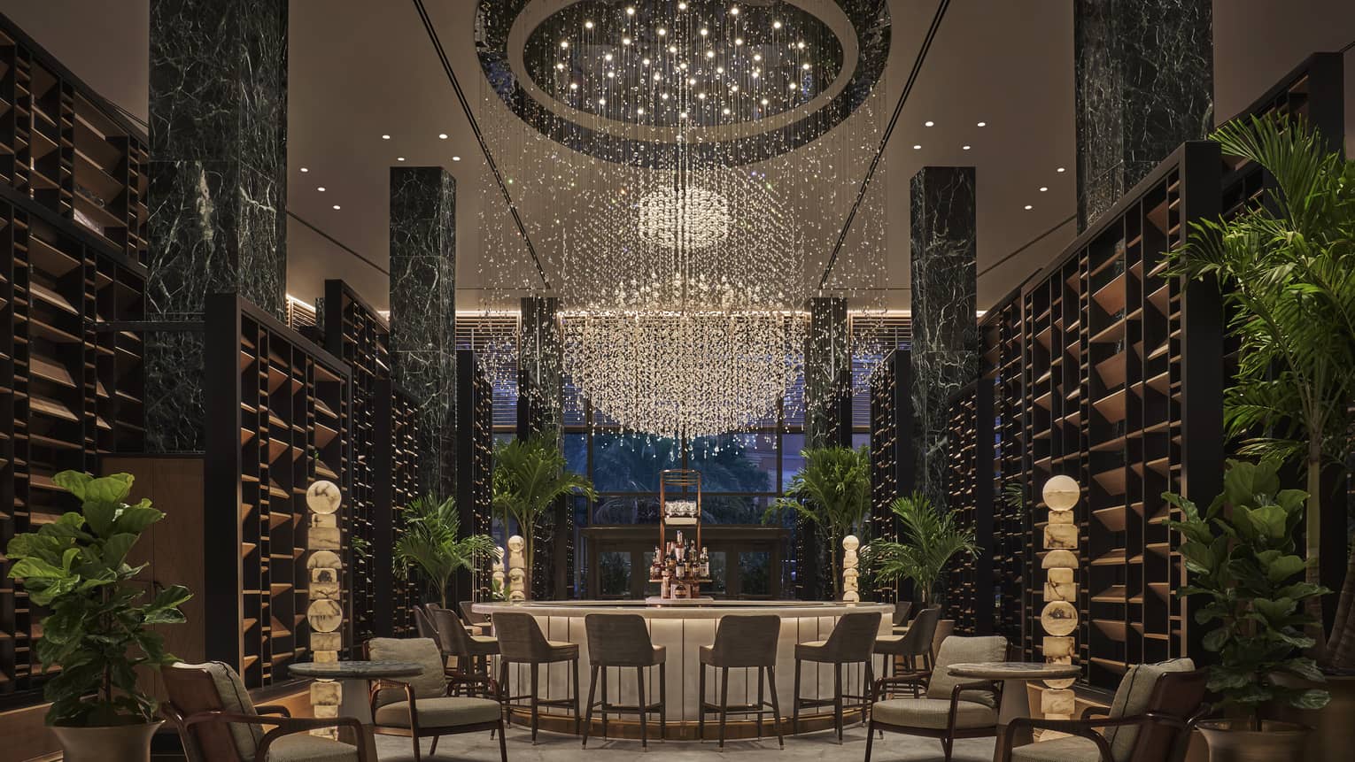 Chandelier Bar with six jade pillars flanked by wooden shelves and potted palms, glass-topped bar in the middle with six cocktail chairs, giant chandelier hanging overhead