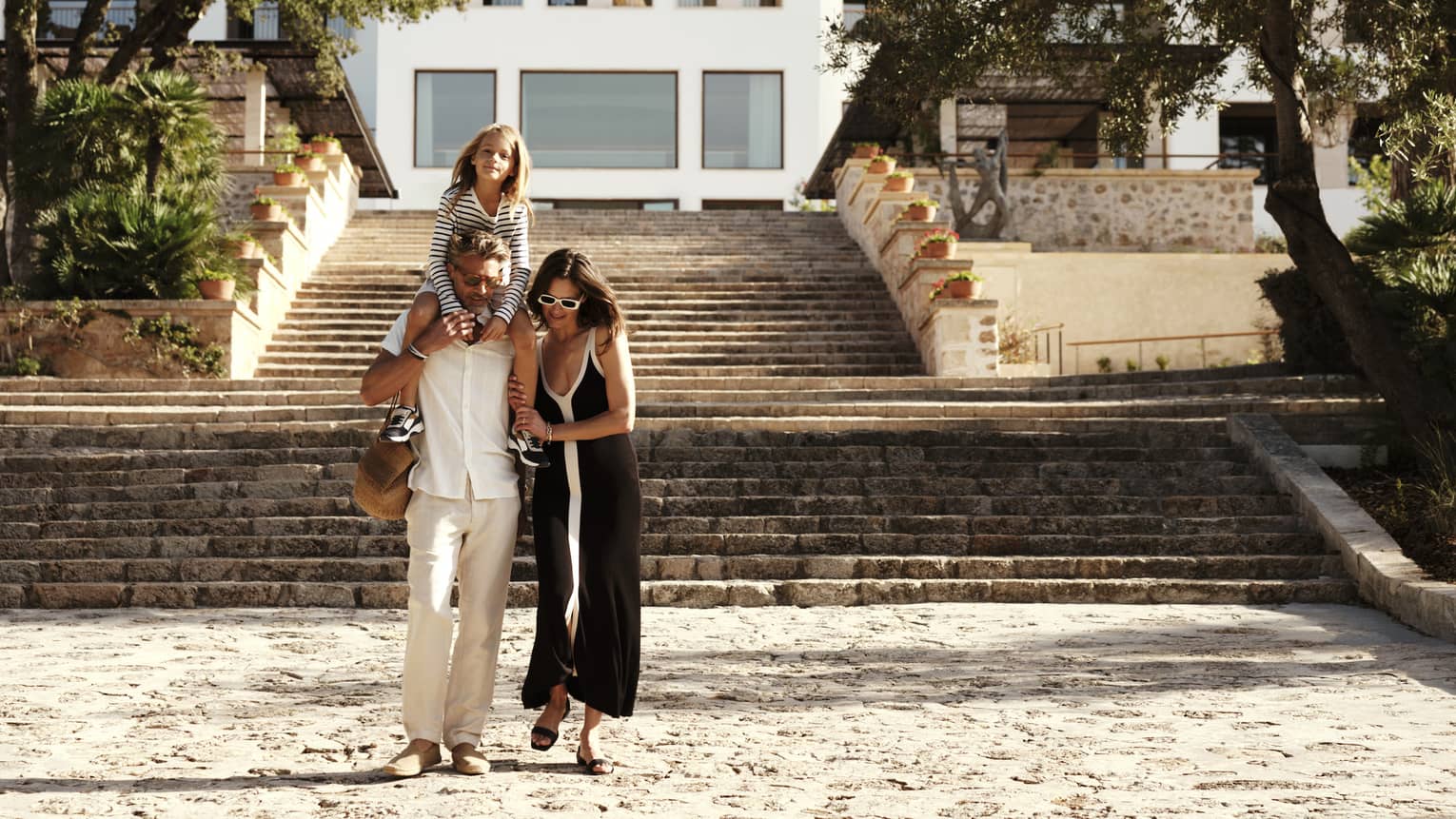 A family strolls in front of a grand stone staircase leading to an elegant white resort at the foot of a lush, treed cliff.