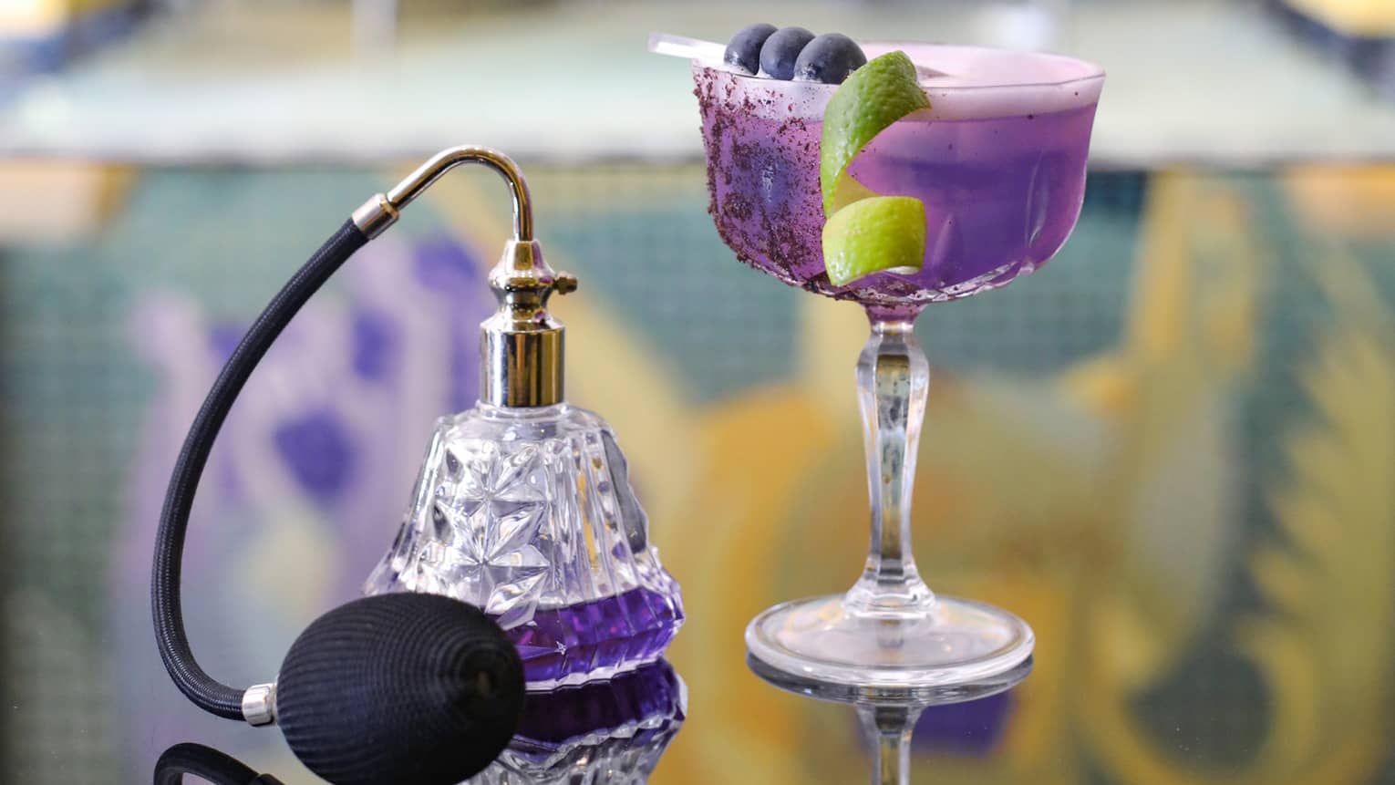 deep purple cocktail in saucer champagne glass garnished with fruit spear and lime twist, beside spray perfume bottle with deep purple liquid inside
