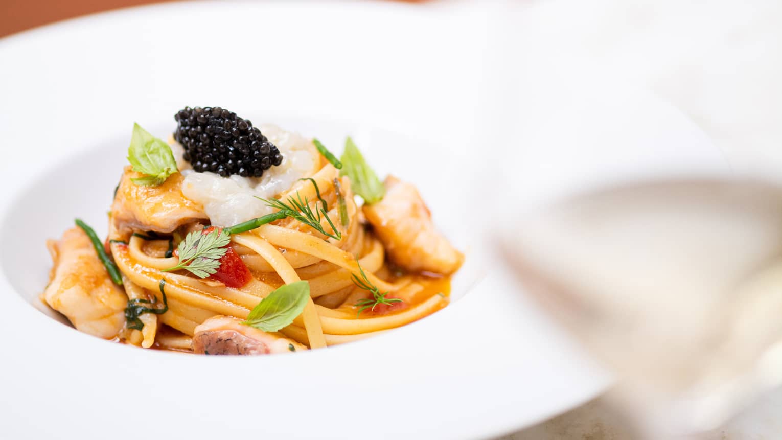 A swirl of seafood linguine topped with black caviar and fresh herbs in a wide pasta bowl, served with a glass of white wine.