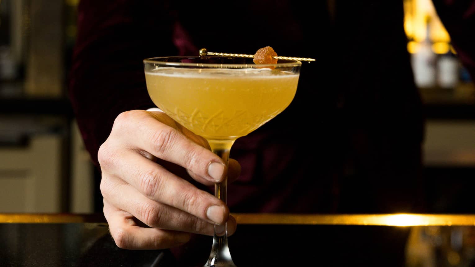 A bartender dressed in a velvet tuxedo serves an orange cocktail at a bar