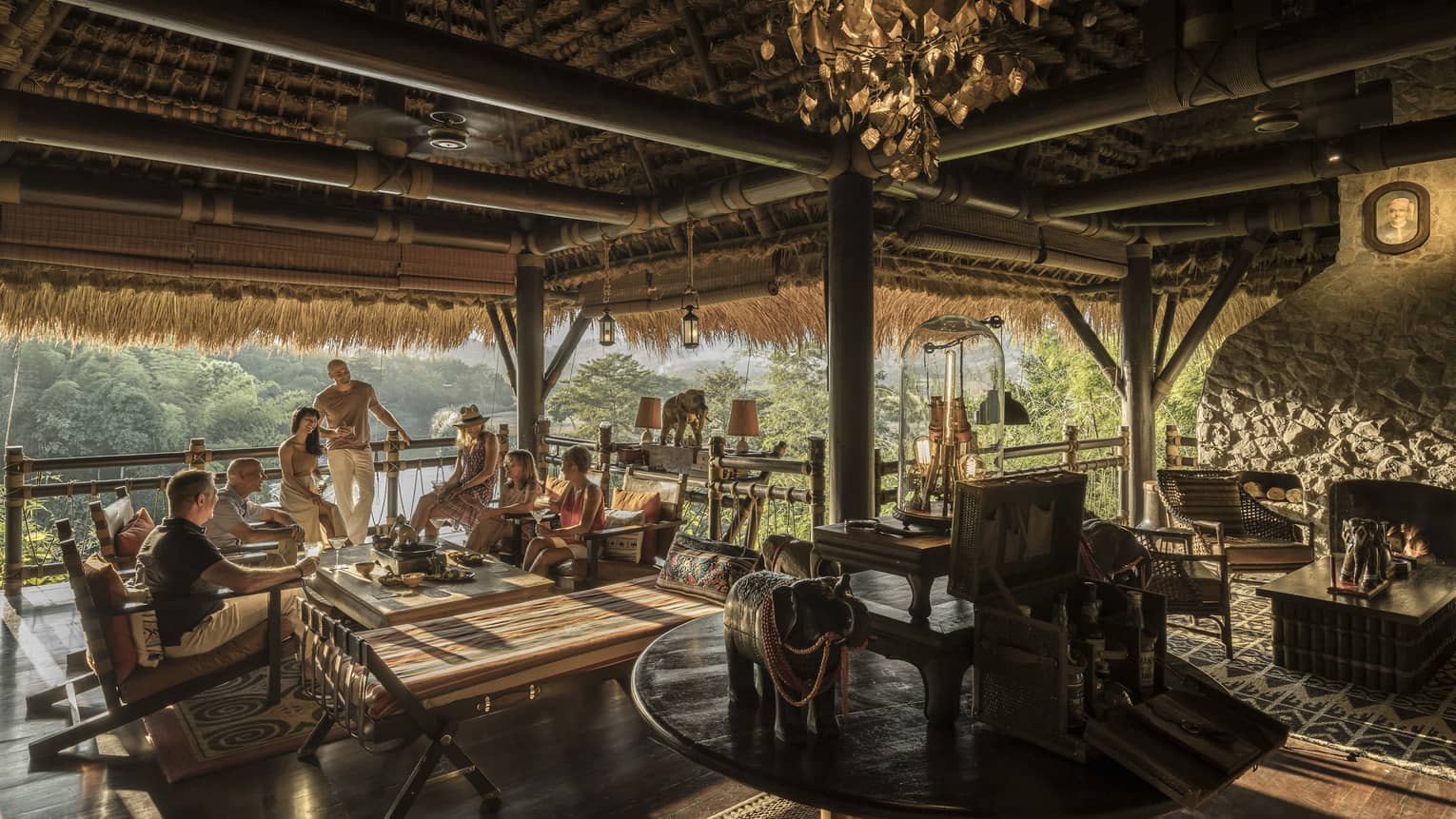 People mingle at open-air bar, beamed thatched roof, lounge seating, trees in the distance