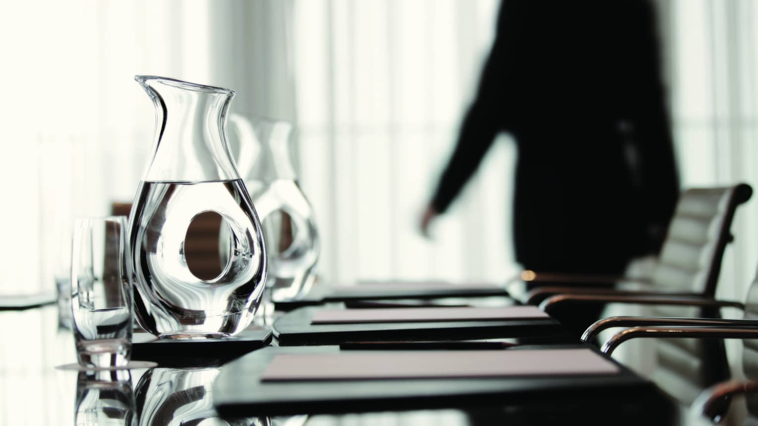 Person wearing business suit walks behind meeting table with agendas, glass water jugs