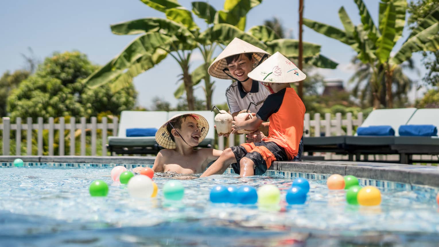 Three laughing children wearing hats, holding coconut drinks play in pool with colourful balls