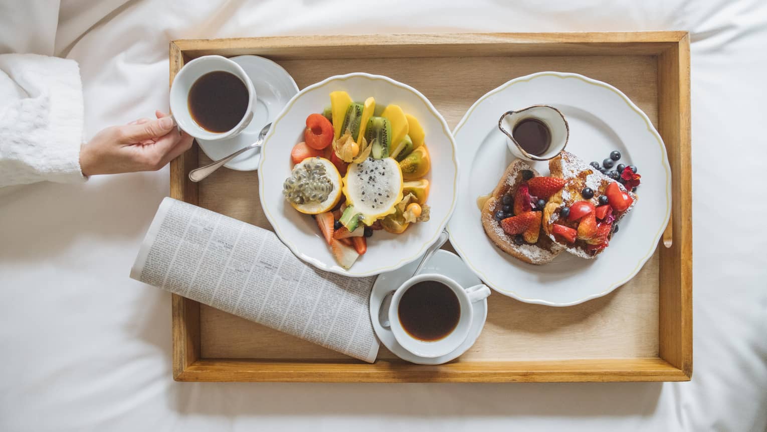 A wooden tray with various fruit, French toast and maple syrup served on white scalloped plates and two cups of coffee 