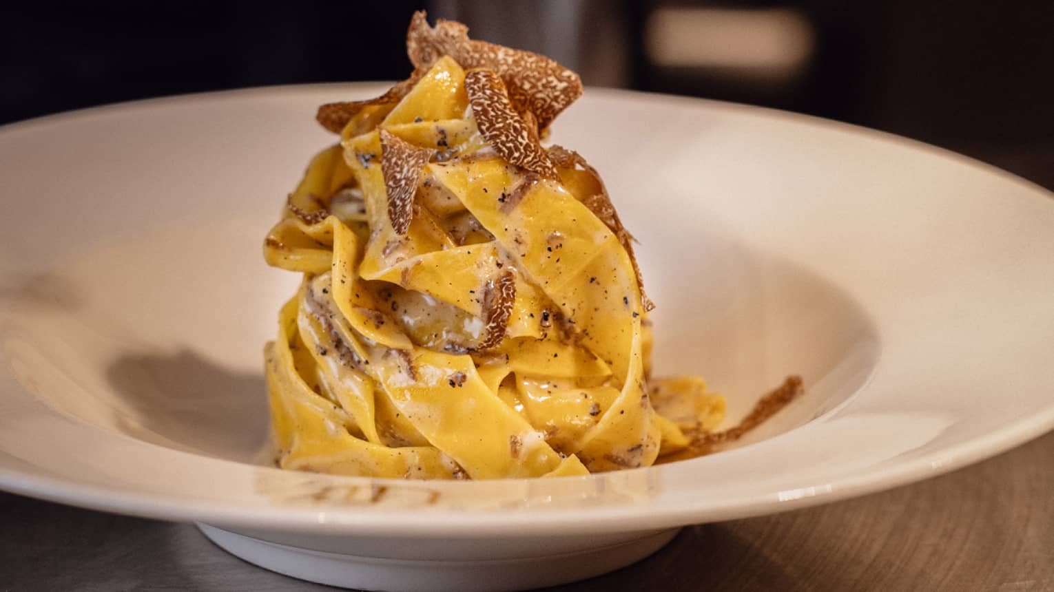 Black truffle shavings fall atop a tall pile of tagliatelle pasta coated in cream sauce, served in a white wide-brimmed bowl.