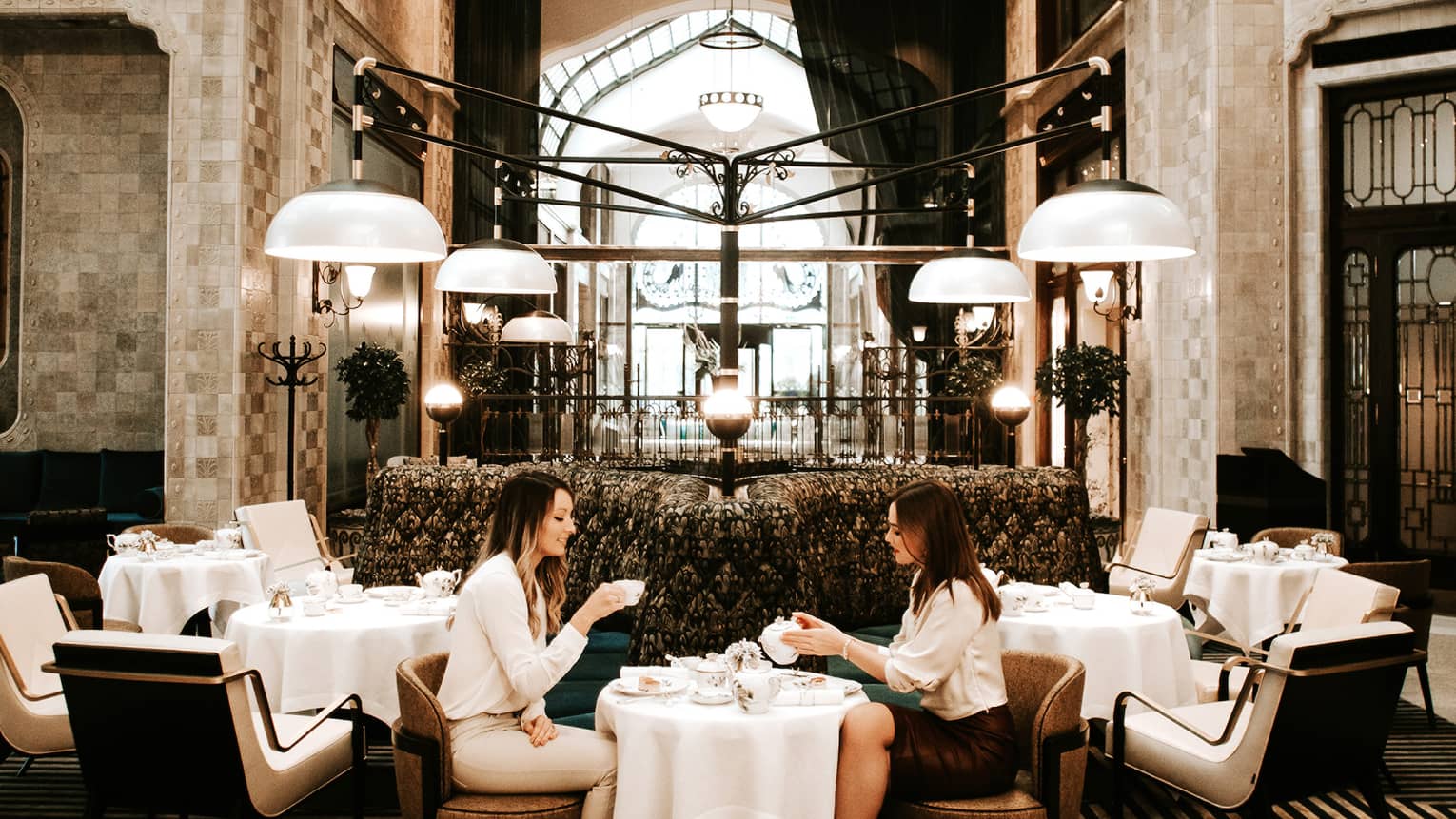 Elegant dining area with a grand chandelier, high arched glass ceiling, and modern seating, where two people are enjoying tea at a small table.