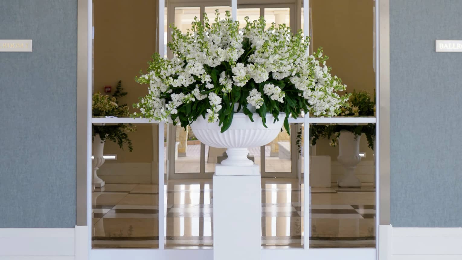 An elegant pedestal with a lush arrangement of white flowers stands in front of a tall arched window set into a slate blue wall.
